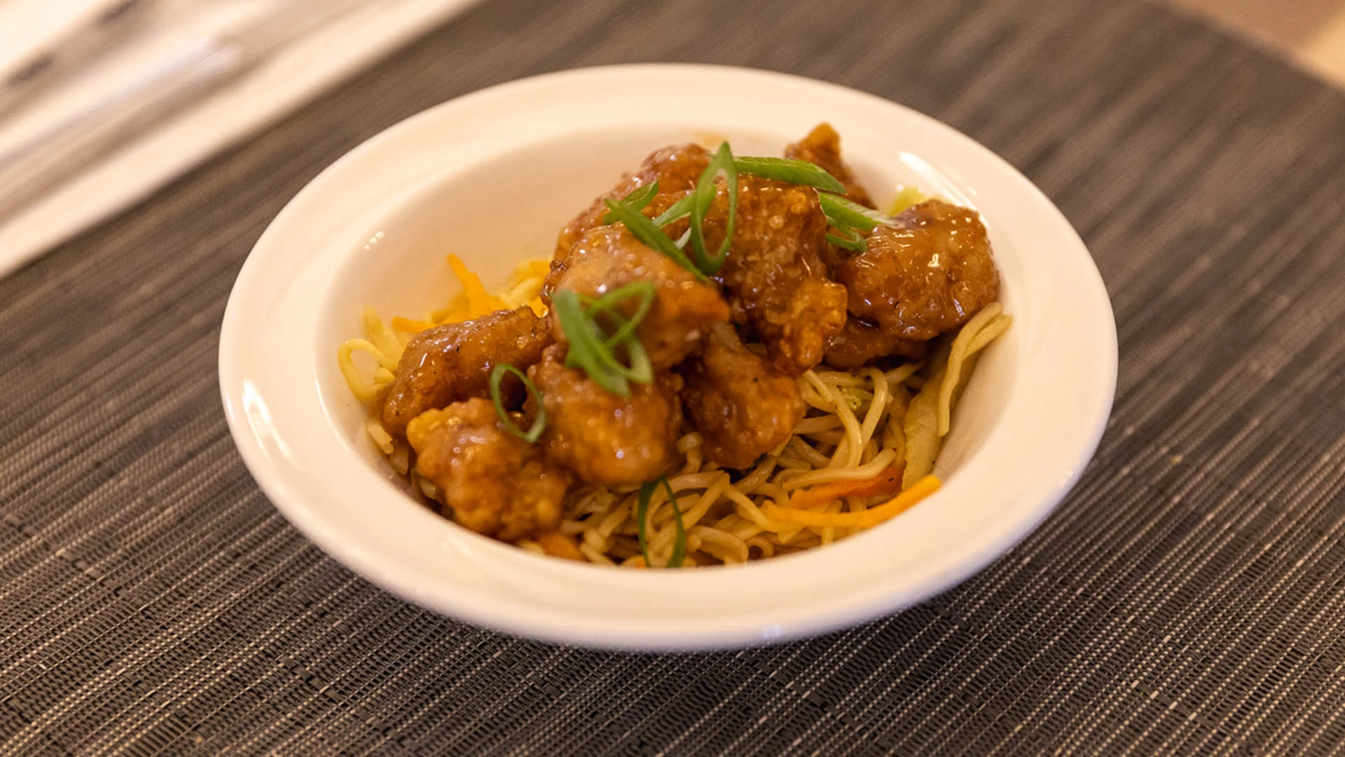 A bowl of orange chicken served over stir-fried noodles and vegetables, garnished with green onions, on a dark textured tablecloth.
